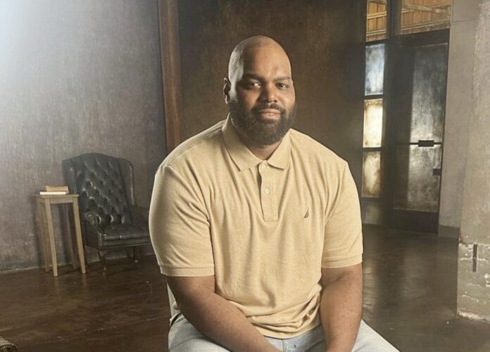 A man sitting indoors wearing a beige polo shirt, smiling gently. A man sitting indoors wearing a beige polo shirt, smiling gently.