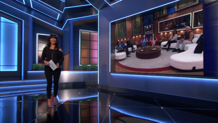 A TV host stands next to a screen showing a panel discussion in a modern studio. A TV host stands next to a screen showing a panel discussion in a modern studio.