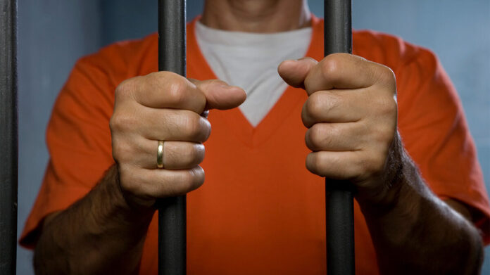 Hands gripping prison bars with a man in an orange jumpsuit behind them. Hands gripping prison bars with a man in an orange jumpsuit behind them.