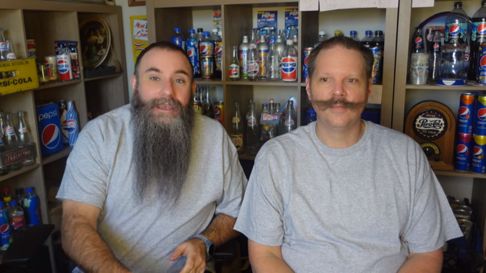Two men with distinctive facial hair sitting indoors, smiling. Two men with distinctive facial hair sitting indoors, smiling.
