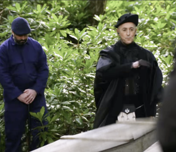 Two individuals in distinctive outfits standing near greenery and a stone structure. Two individuals in distinctive outfits standing near greenery and a stone structure.