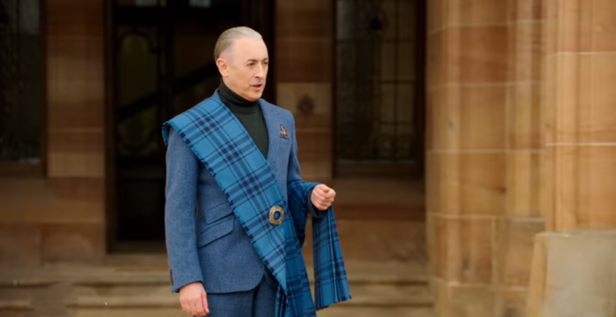 Man in a blue suit and tartan sash stands confidently. Man in a blue suit and tartan sash stands confidently.