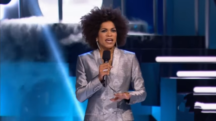 A woman passionately speaking on stage in a shiny silver suit, holding a microphone. A woman passionately speaking on stage in a shiny silver suit, holding a microphone.