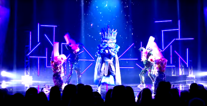 A performer in an elaborate costume and crown on stage with dancers. A performer in an elaborate costume and crown on stage with dancers.