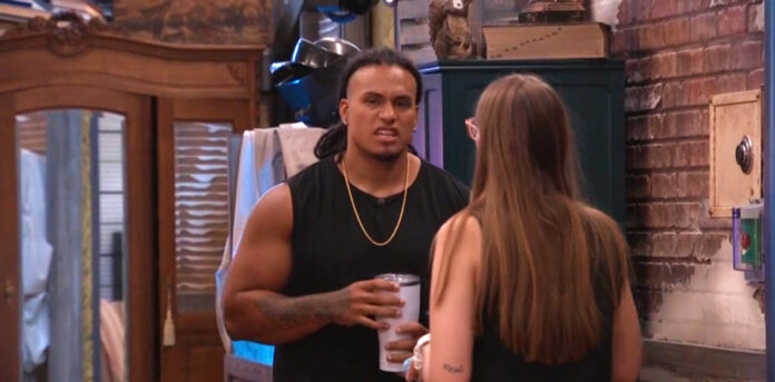 A man in a black sleeveless shirt holding a cup, talking to a woman with long hair indoors. A man in a black sleeveless shirt holding a cup, talking to a woman with long hair indoors.