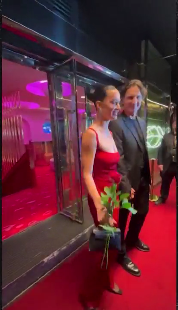A woman in a white dress and a man in a black suit smile on a red carpet.