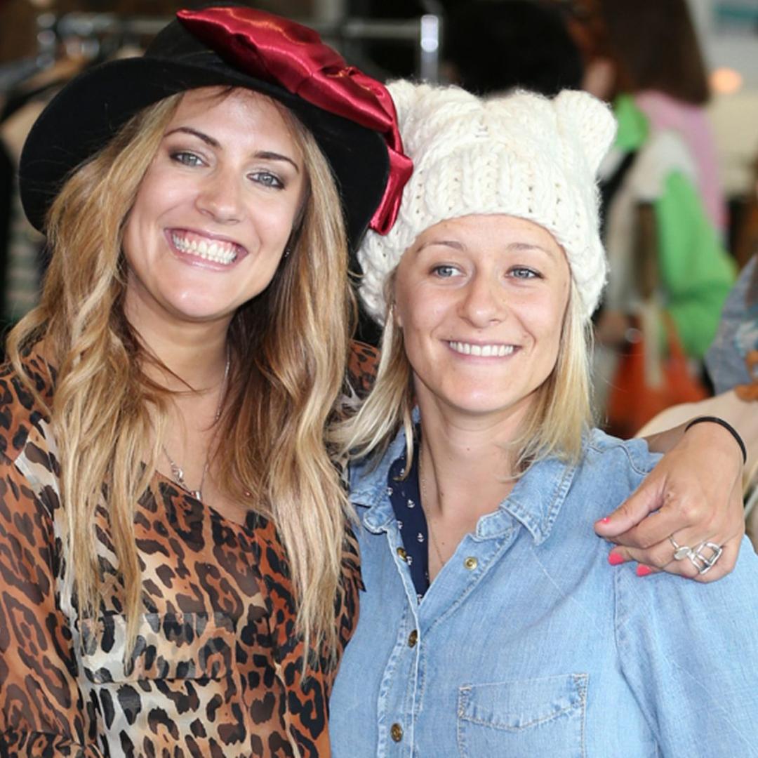 Two women smiling, one in a leopard print top and black hat, the other in a light blue shirt and knitted white hat.